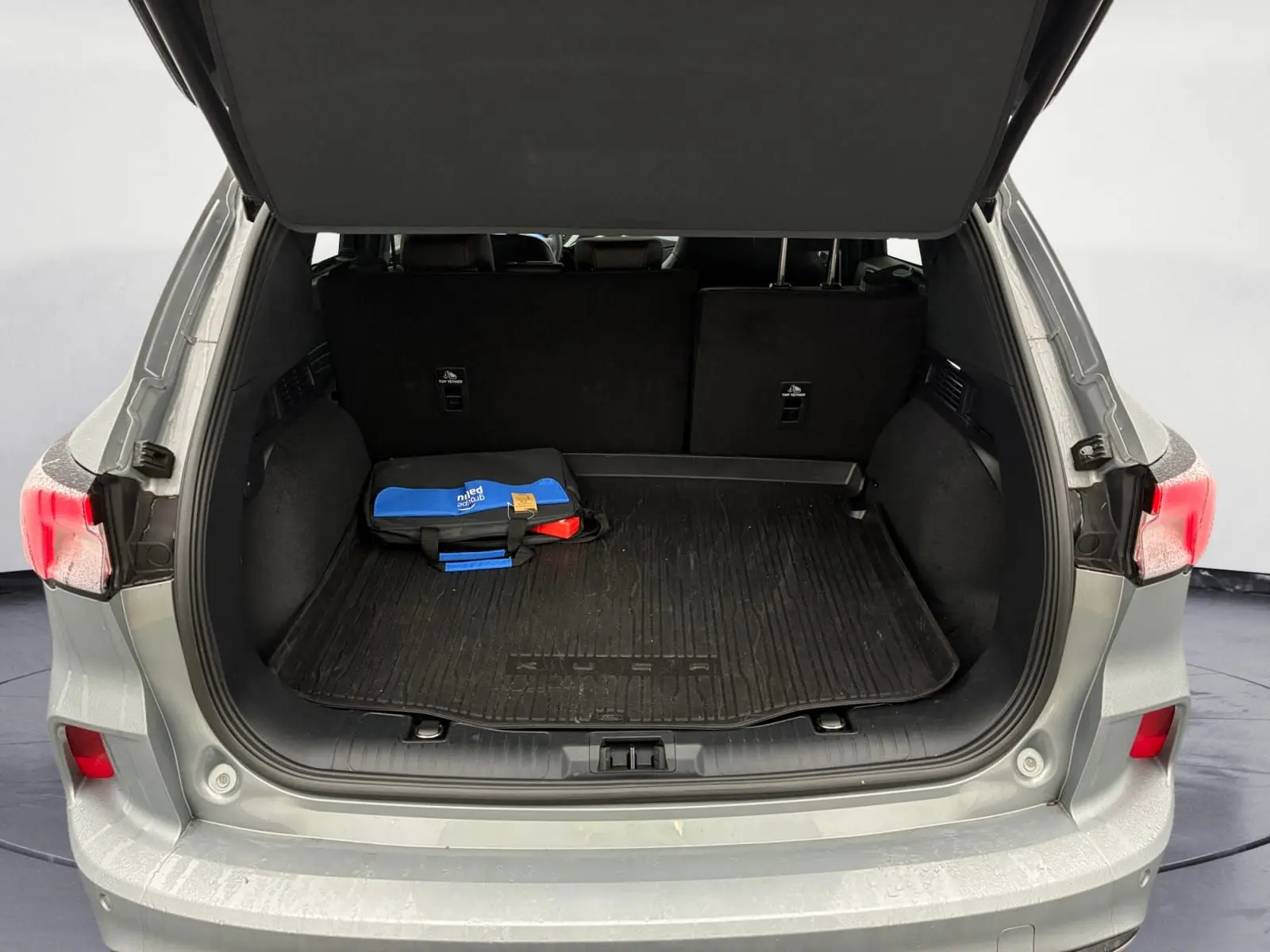 Coffre ouvert vu de l'arrière d'un Ford Kuga gris 2024, avec tapis noir et trousse de secours bleue visible.