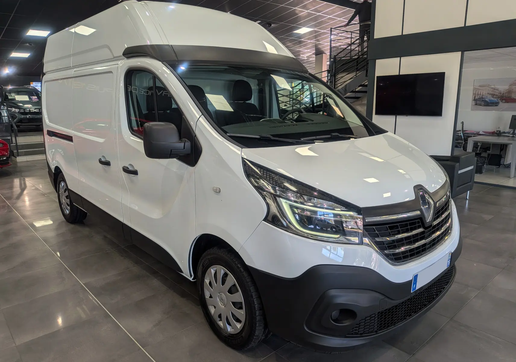 Vue 3/4 avant d'un Renault Trafic Fourgon blanc Glacier L2H2, avec calandre noire et phares LED dans un showroom.