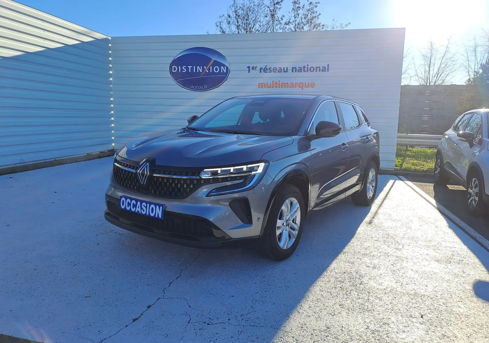 Renault Austral gris schiste en 3/4 avant droit, avec calandre distinctive et feux LED allumés, sur parking extérieur.