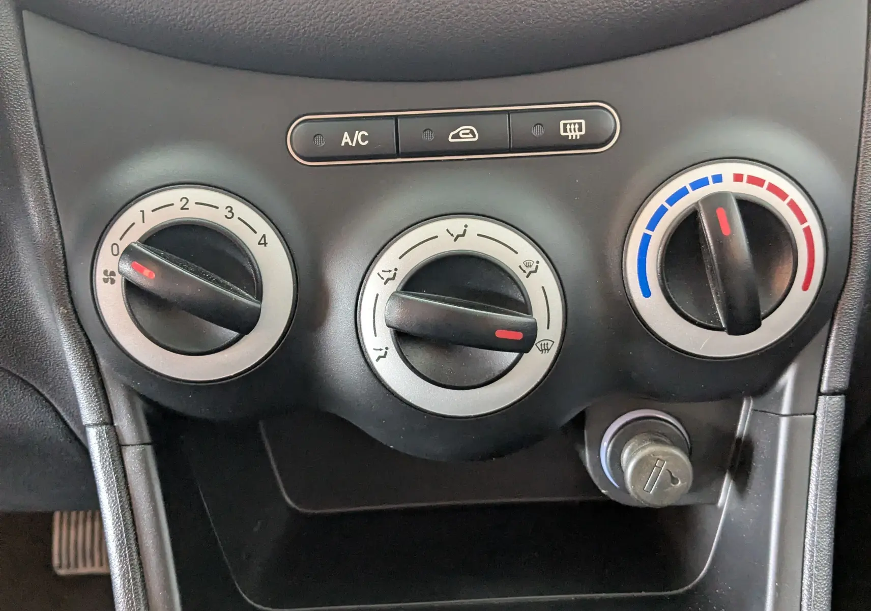 Gros plan sur les commandes de climatisation et ventilation d'une Hyundai i10 gris clair de 2010, tableau de bord noir.