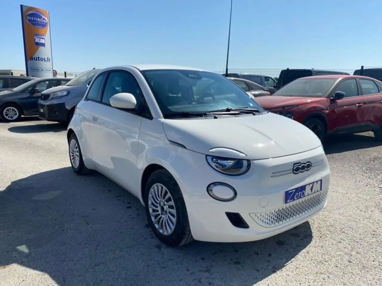 Vue 3/4 avant droite d'une Fiat 500 électrique blanche métallisée 2022 avec logo 500 et plaque Zero KM visible.