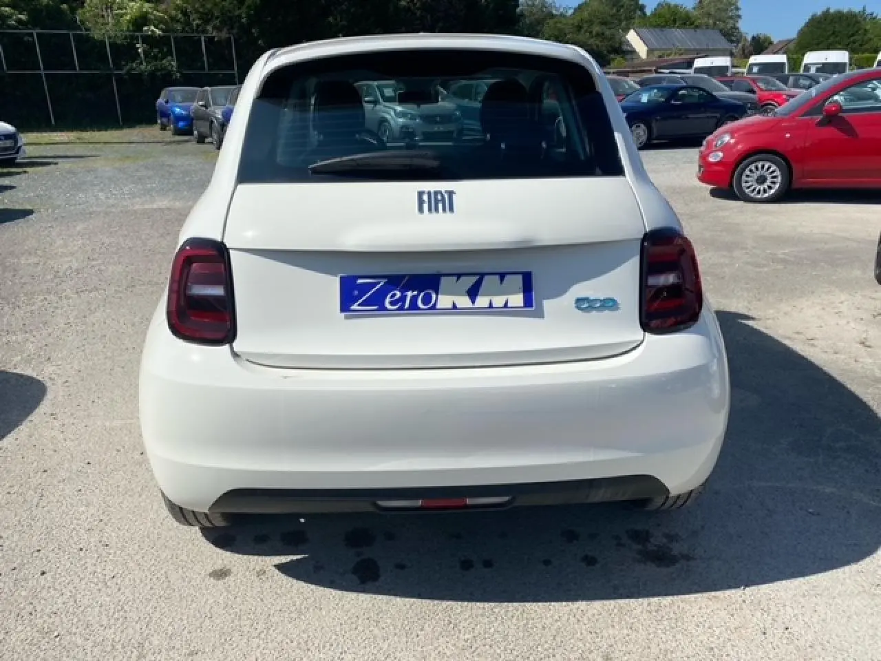 Vue arrière d'une FIAT 500 électrique blanche 2022 avec logo bleu et plaque "Zero KM" sur un parking extérieur.