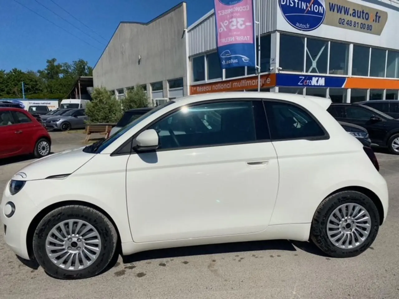 Vue de profil côté gauche d'une FIAT 500 électrique blanche métallisée, stationnée devant un bâtiment commercial.