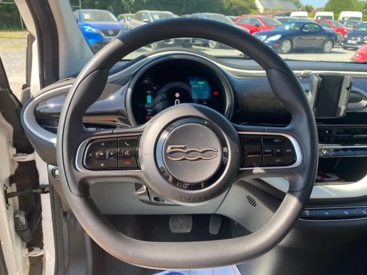 Vue intérieure centrée sur le volant noir de la Fiat 500 électrique blanche, avec tableau de bord numérique et support smartphone visible.