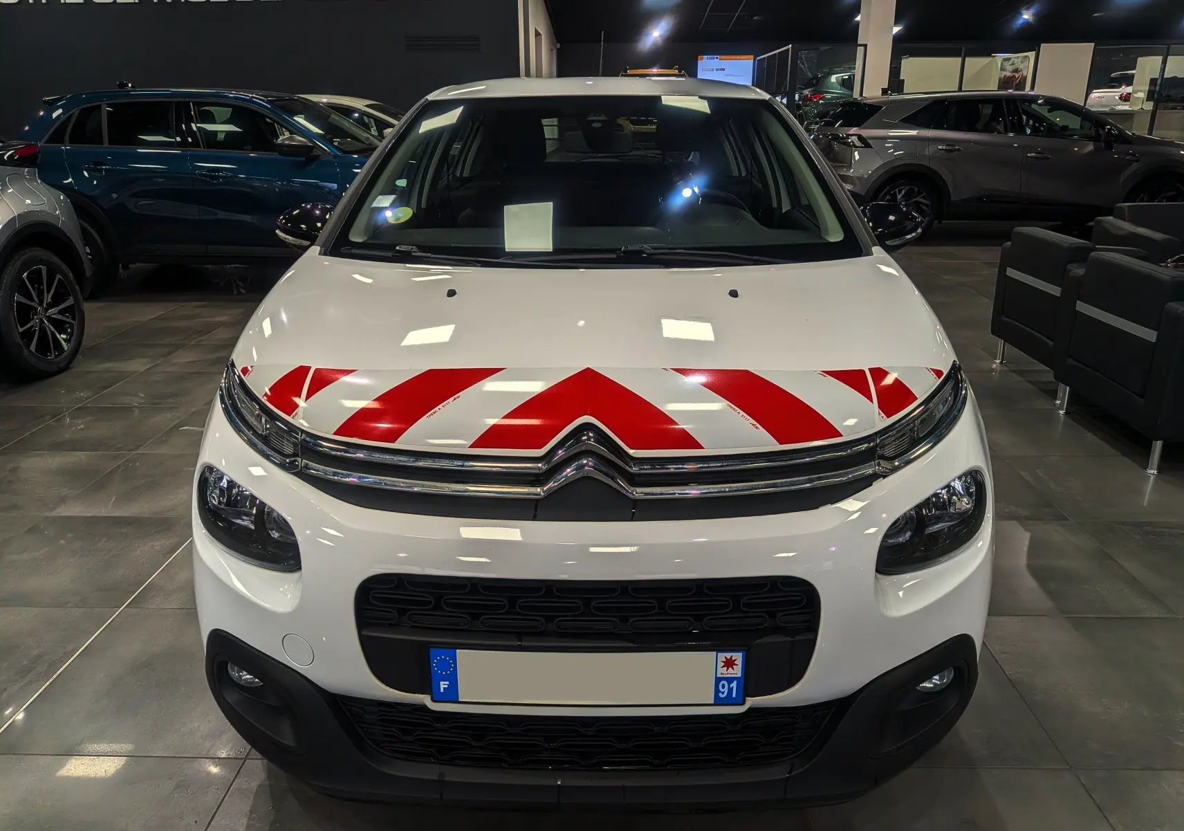 Vue frontale d'une Citroën C3 Société blanche avec bandes rouges de signalisation sur le capot, en showroom.