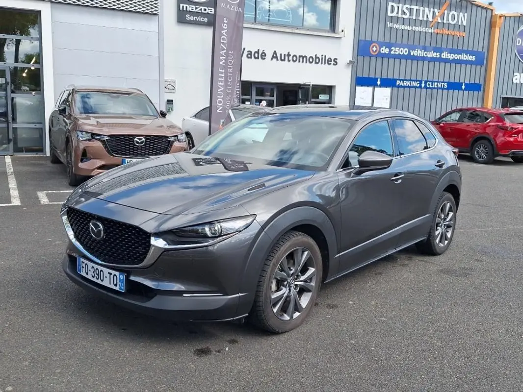 Mazda CX-30 gris en 3/4 avant droit sur parking devant concession avec jantes aluminium visibles.