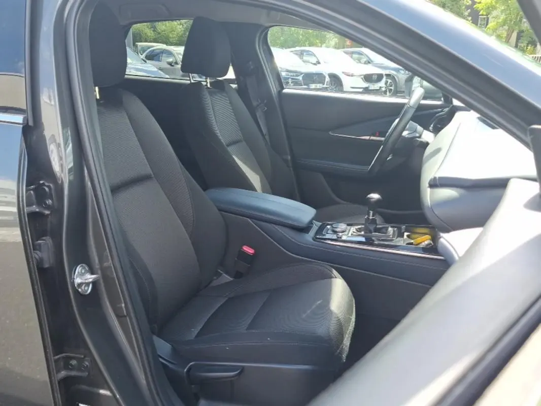 Vue intérieure côté conducteur du Mazda CX-30 gris, montrant les sièges noirs, accoudoir central et volant multifonctions.