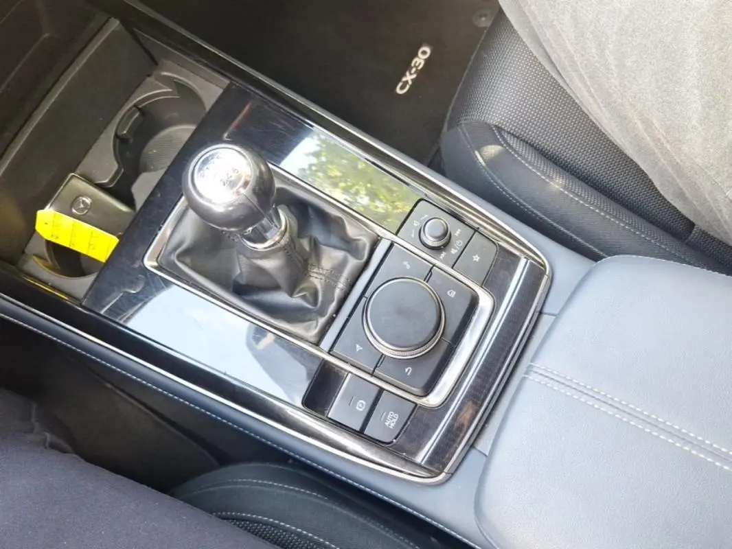 Vue plongeante sur la console centrale noire brillante du Mazda CX-30 avec levier de vitesse manuel et accoudoir gris.