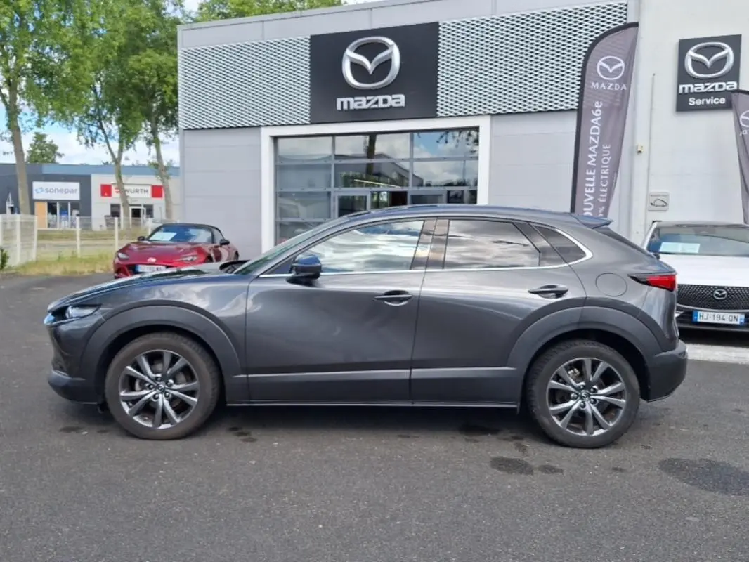 Vue de profil côté gauche d'un Mazda CX-30 gris stationné devant un concessionnaire Mazda avec jantes aluminium.