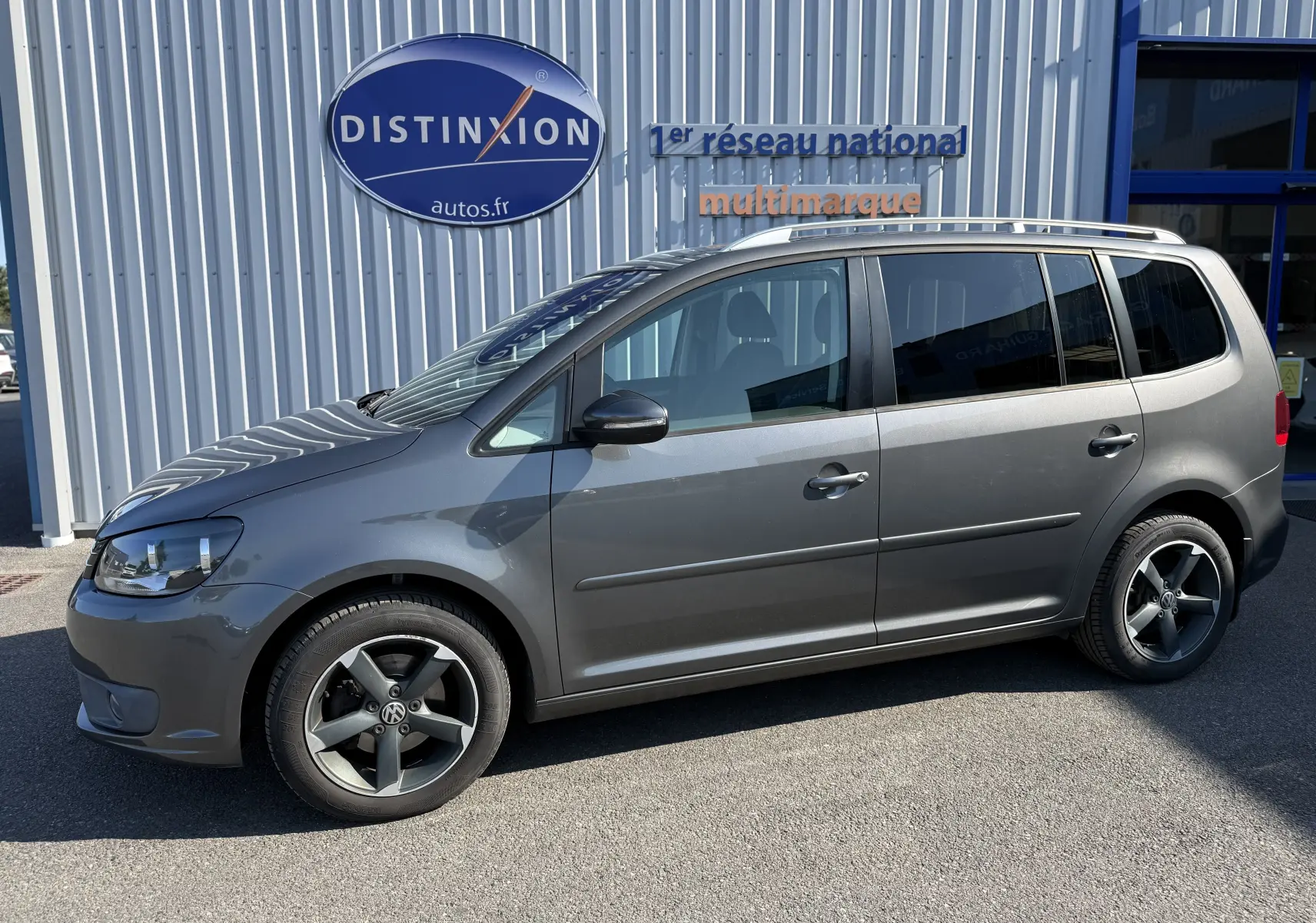 Vue de profil côté gauche d’un Volkswagen Touran gris 2012 avec jantes alliage noires et toit ouvrant visible.
