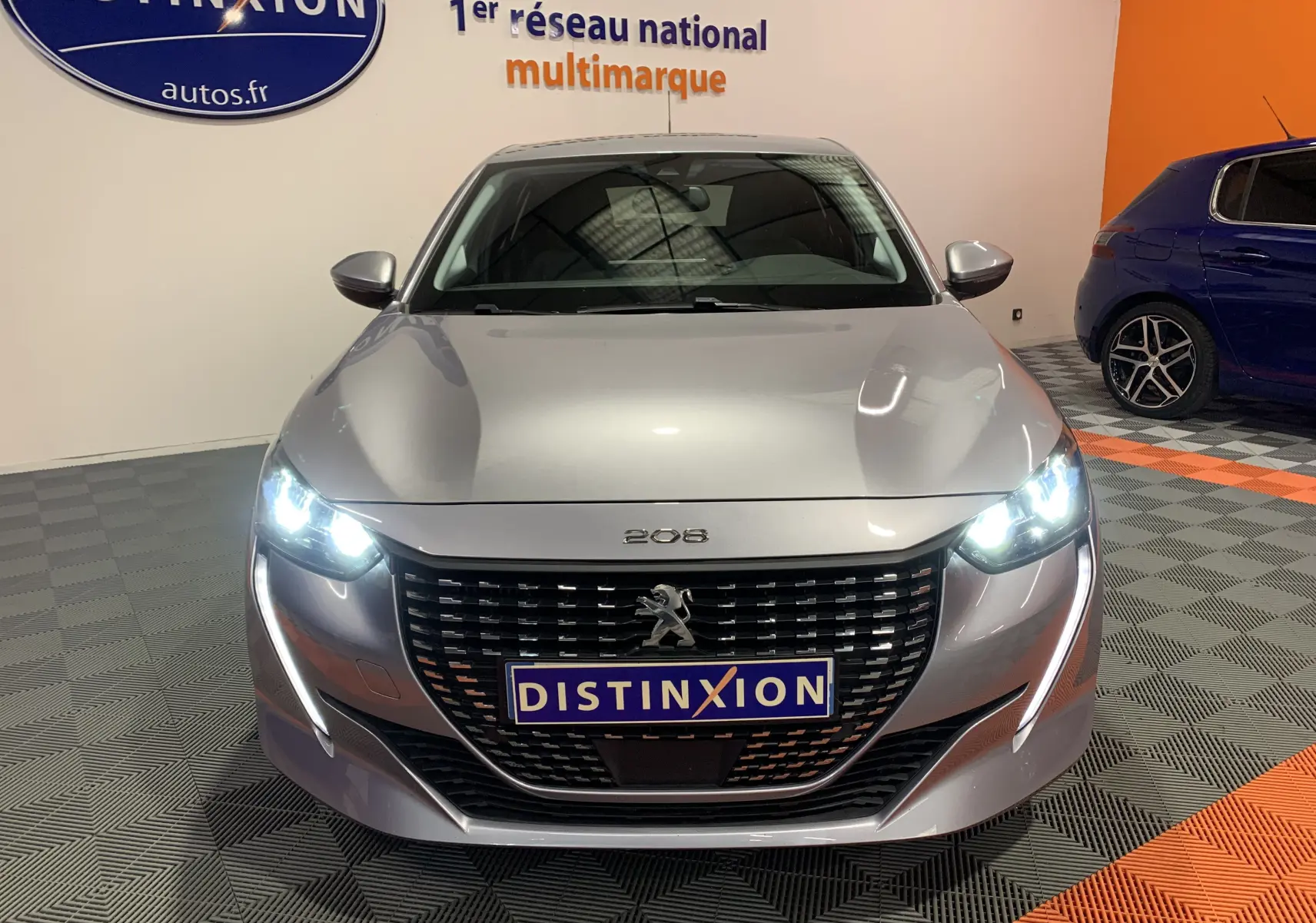 Vue frontale d'une Peugeot 208 gris métallisé avec phares allumés dans un showroom multimarque.