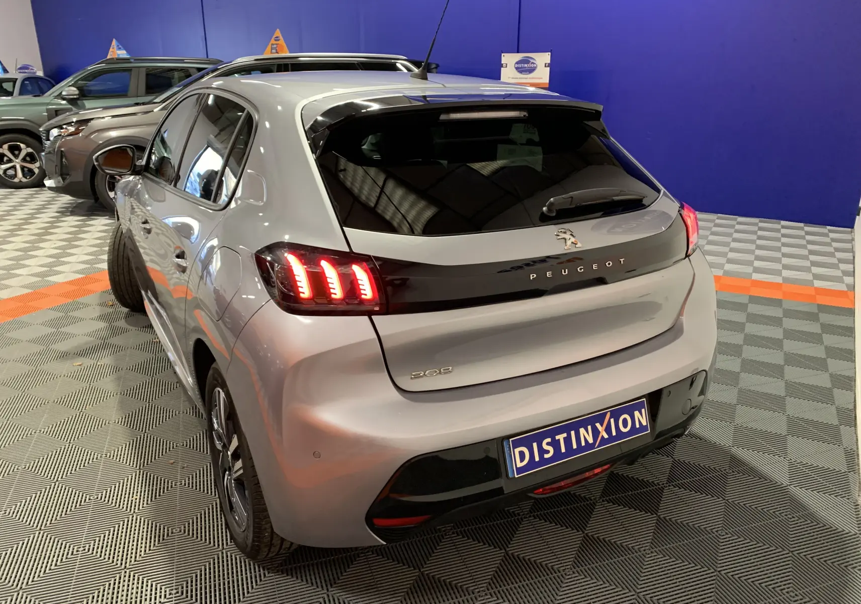 Vue 3/4 arrière droite d'une Peugeot 208 gris clair avec feux arrière LED allumés dans un showroom.