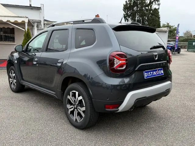Vue 3/4 arrière droite d'un Dacia Duster gris comète 2022 avec feux arrière LED et barres de toit noires.