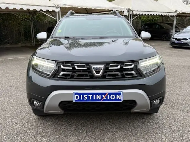 Vue avant d'un Dacia Duster 2022 gris comète avec calandre noire et feux LED allumés sur un parking.