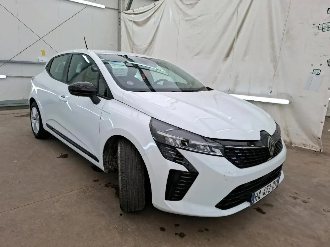 Renault Clio blanche vue 3/4 avant droit, avec calandre noire et feux LED distinctifs, stationnée en intérieur.