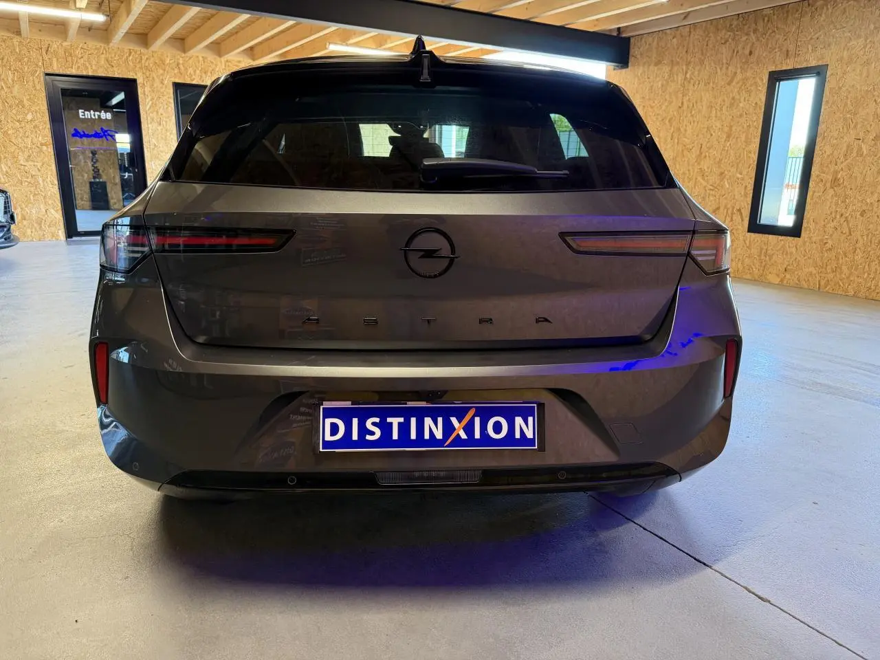 Vue arrière d'une Opel Astra gris foncé avec logo noir et pare-chocs arrière noir dans un garage lumineux.