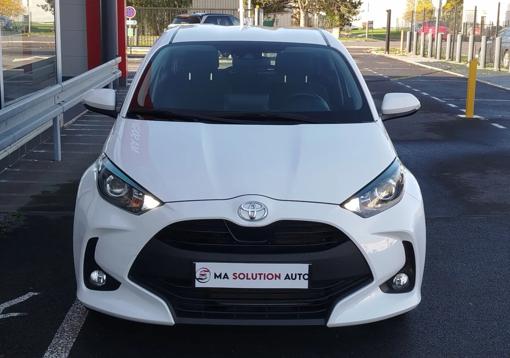 Vue avant d'une Toyota Yaris blanche pur 2023, avec calandre noire et phares allumés, stationnée devant un bâtiment.