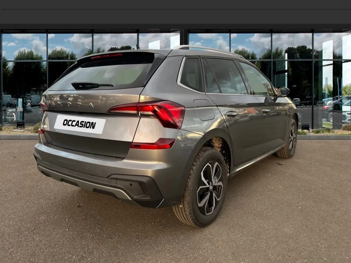 Vue 3/4 arrière droite d'un Skoda Kamiq gris clair avec jantes noires et toit ouvrant, stationné devant une vitrine.