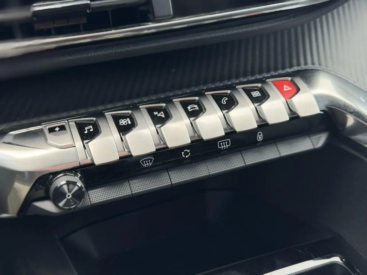 Gros plan sur la rangée de boutons métalliques du tableau de bord du Peugeot 3008 Plug-in Hybrid noir, avec touches fonctionnelles et alerte.