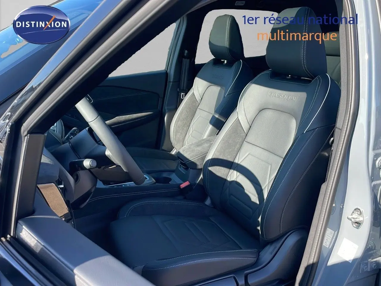 Vue intérieure côté conducteur du Nissan Qashqai gris céramique, sièges cuir noir avec logo Qashqai brodé et toit panoramique ouvert.