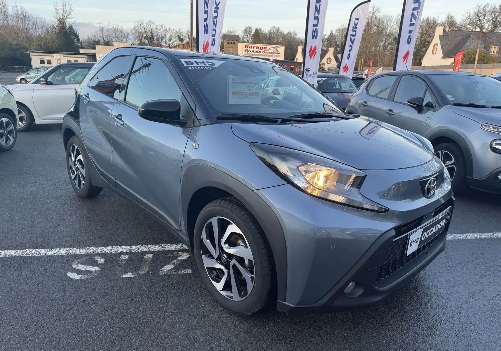 Toyota Aygo X bleu vue 3/4 avant droit avec phares allumés et jantes alliage distinctives sur parking.