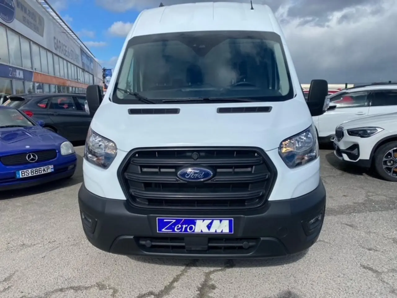 Vue avant d'un Ford Transit Fourgon blanc avec calandre noire et plaque d'immatriculation "Zéro KM" sur un parking.