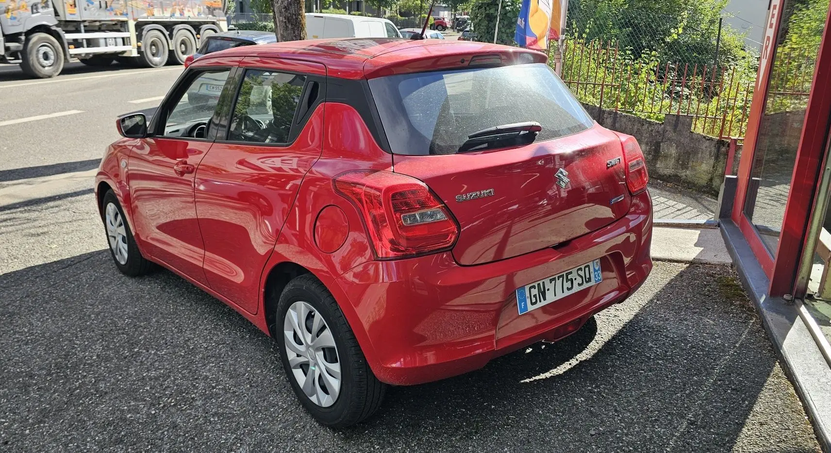 Vue 3/4 arrière droite d'une Suzuki Swift 1.2 Dualjet Hybrid 2023 en rouge Fervent, avec hayon et feux arrière visibles.