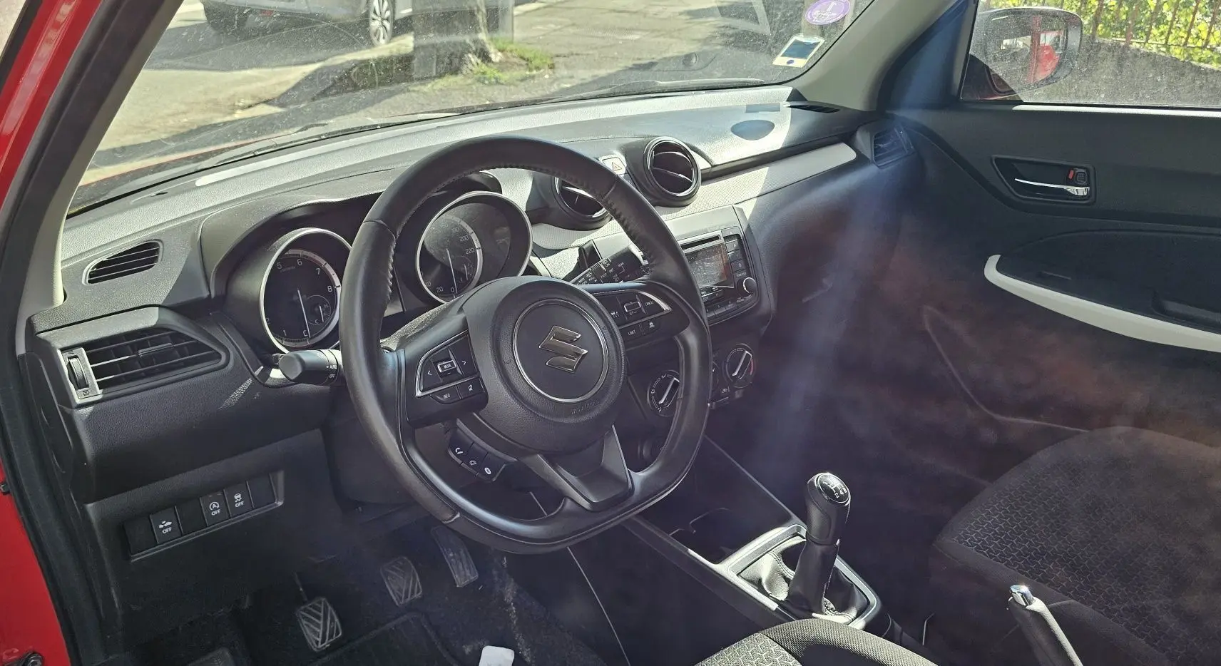 Intérieur de la Suzuki Swift 1.2 Dualjet Hybrid 2023 en rouge, vue côté conducteur avec volant et tableau de bord visibles.