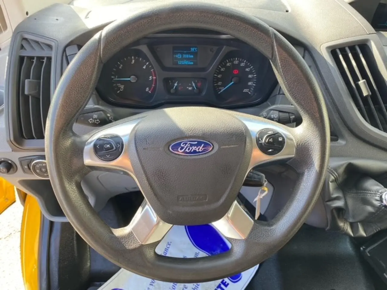 Vue rapprochée du volant Ford noir avec commandes intégrées, tableau de bord et levier de vitesse du Transit Fourgon orange 2015.