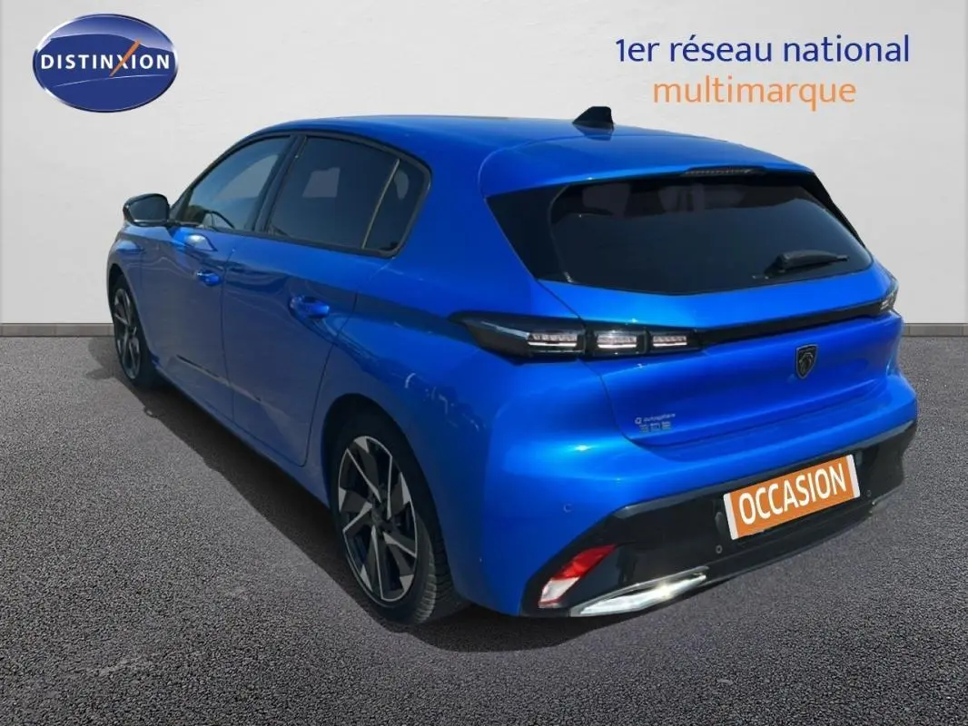 Vue 3/4 arrière gauche d'une Peugeot 308 bleu clair avec jantes alliage et vitres teintées, stationnée sur sol gris.