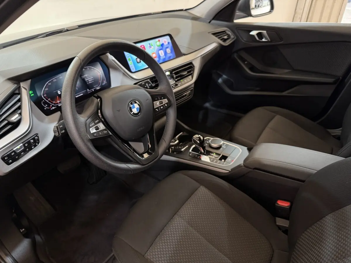 Vue intérieure côté conducteur de la BMW Série 1 116i 2023, tableau de bord moderne avec écran tactile et volant cuir noir.