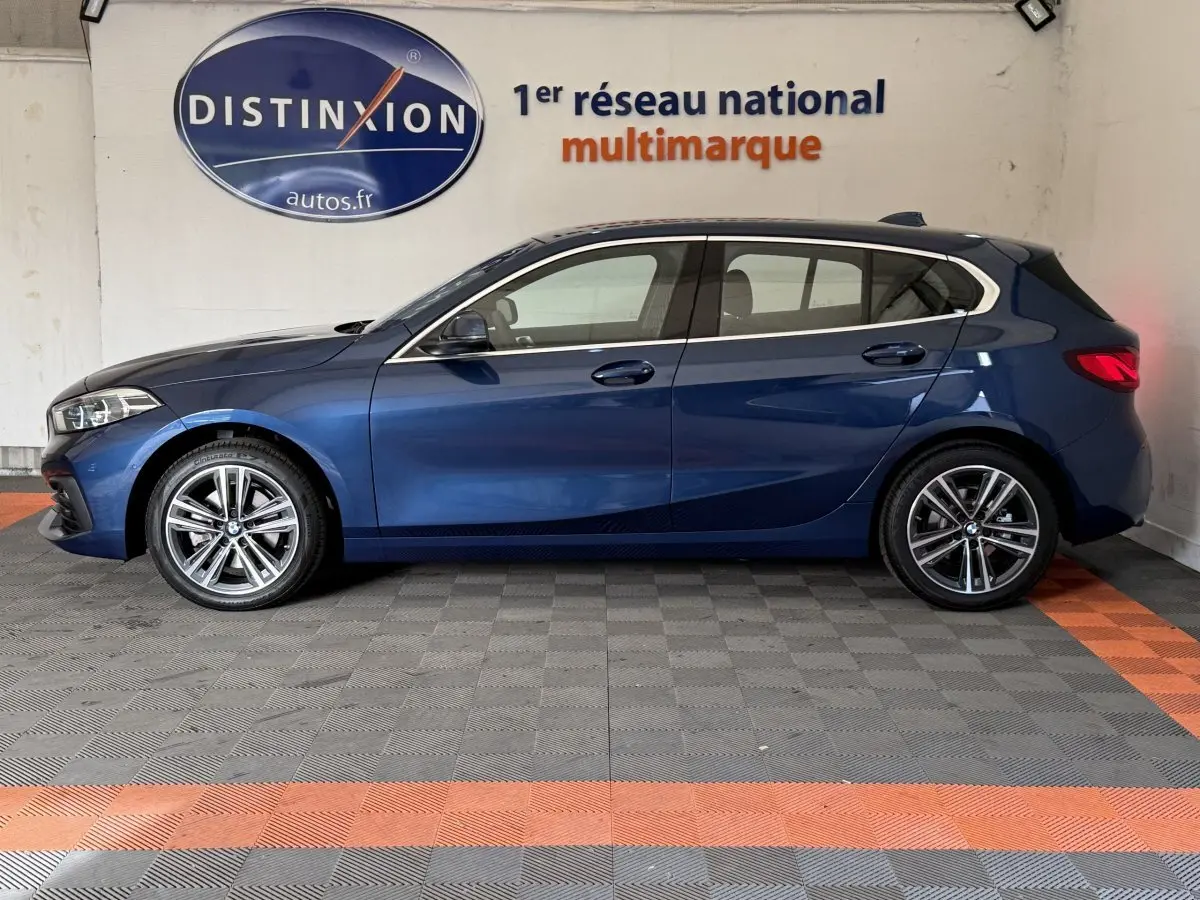 BMW Série 1 116i Business Design bleu vue de profil côté gauche dans un showroom avec sol gris et orange.