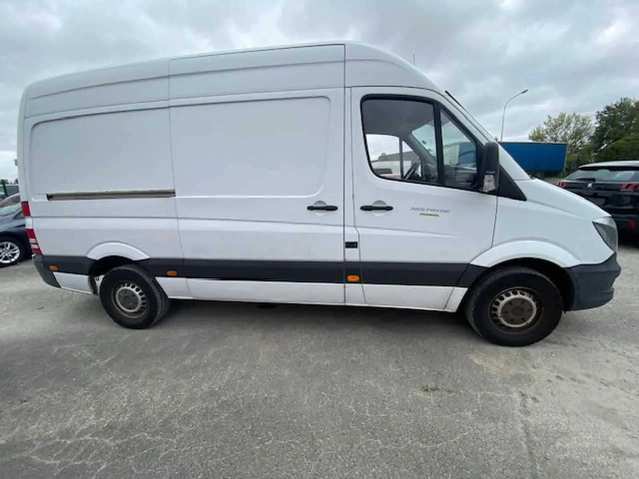 Vue latérale gauche d'un fourgon Mercedes Sprinter blanc de 2015 avec bande noire et jantes acier simples.