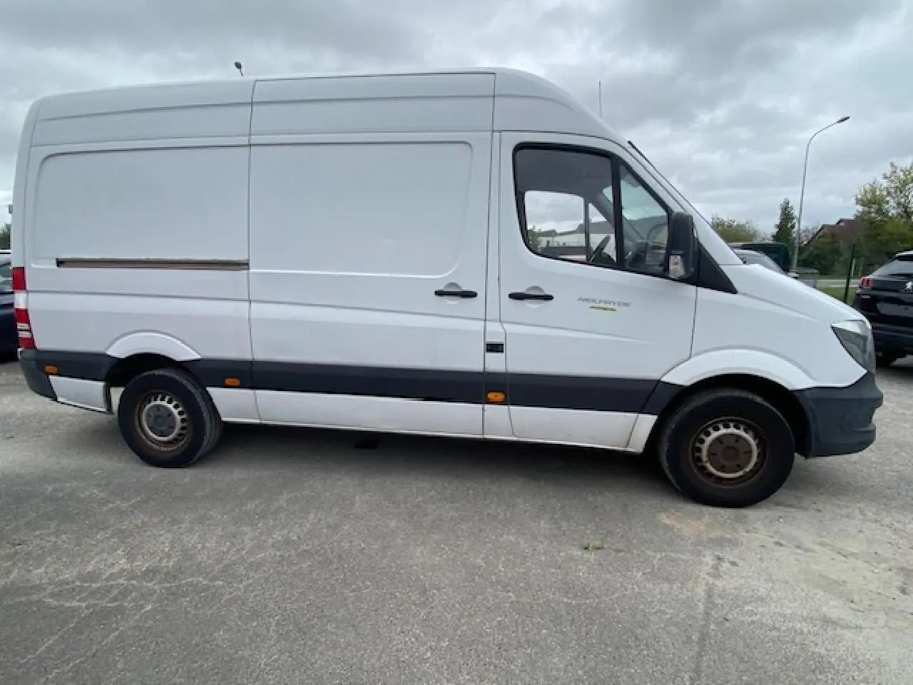 Vue de profil côté gauche d’un Mercedes Sprinter Fourgon blanc de 2015 avec bande noire et feux latéraux orange.