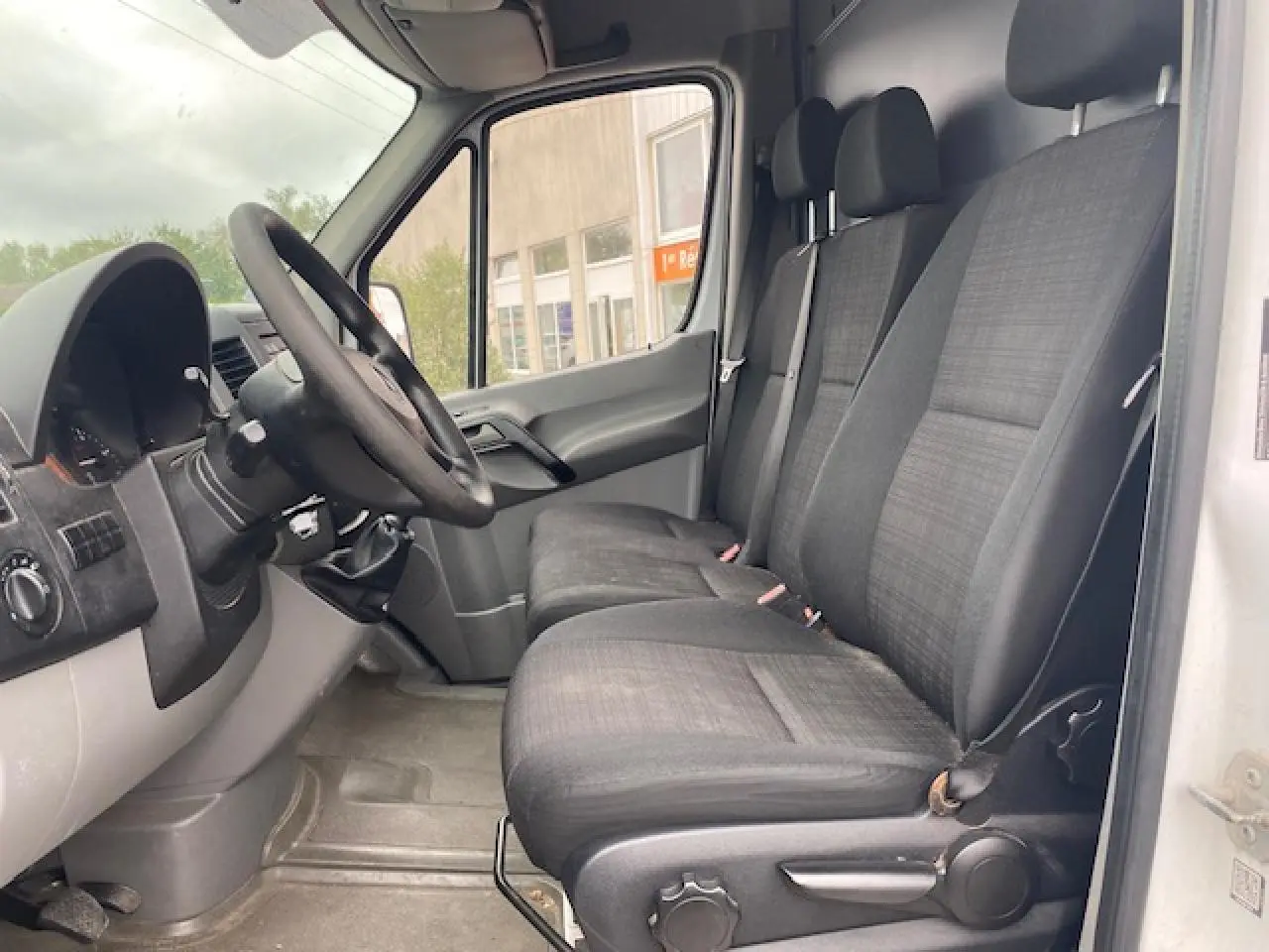 Vue intérieure côté conducteur du Mercedes Sprinter blanc 2015, montrant les sièges avant en tissu gris et le tableau de bord.