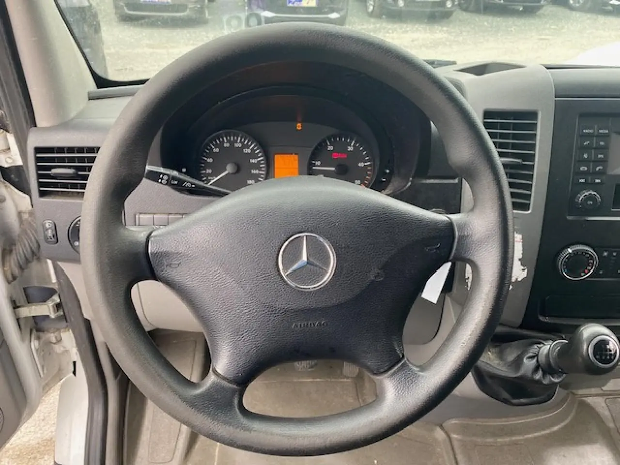 Vue intérieure centrée sur le volant noir Mercedes du Sprinter Fourgon blanc, avec tableau de bord et levier de vitesses manuelle visibles.