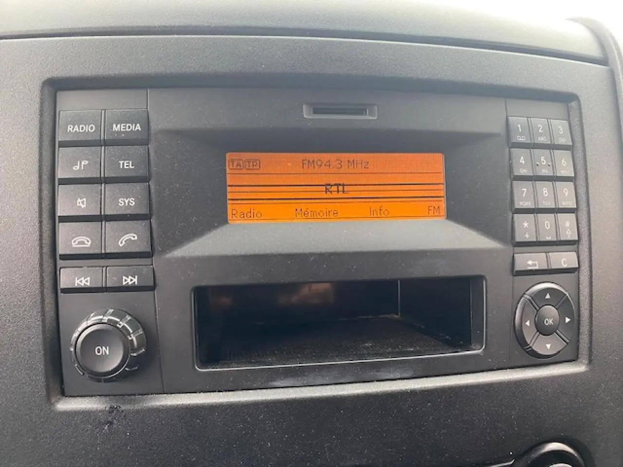 Gros plan sur l’autoradio noir avec écran orange affichant la fréquence FM 94.3 dans un Mercedes Sprinter blanc de 2015.