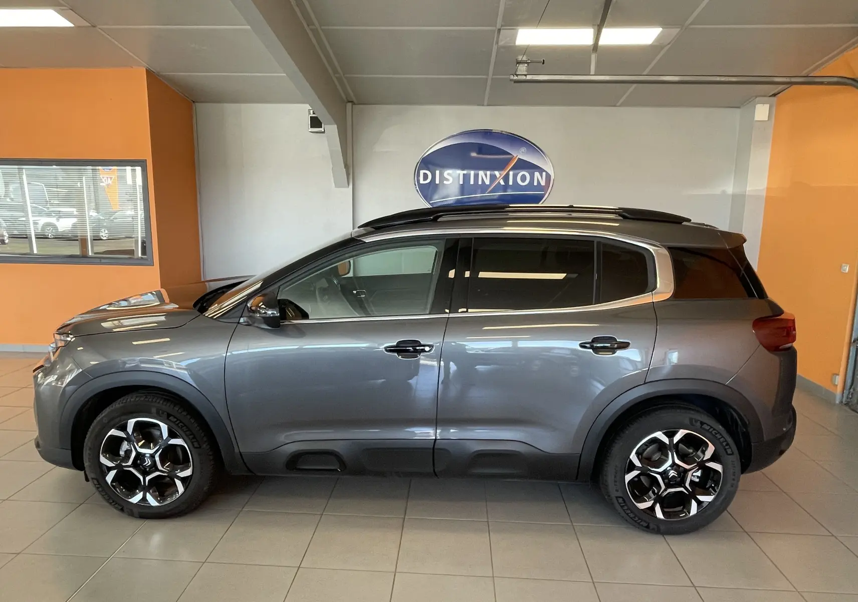 Vue latérale droite d'un Citroën C5 Aircross hybride gris platinium avec jantes alliage biton et barres de toit en intérieur showroom.