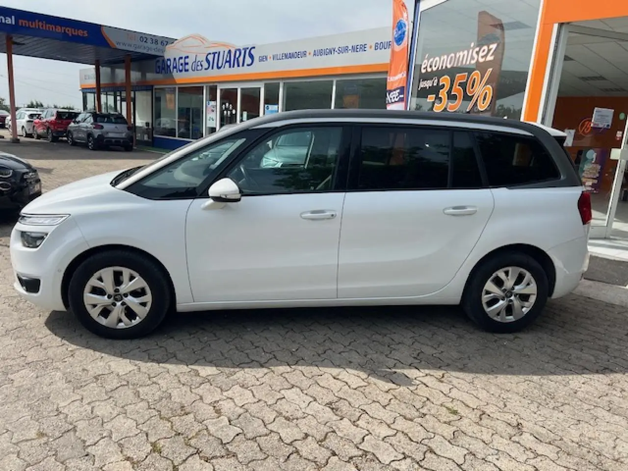 Vue de profil côté gauche d'un Citroën Grand C4 Picasso blanc stationné devant un garage avec vitres latérales surteintées.