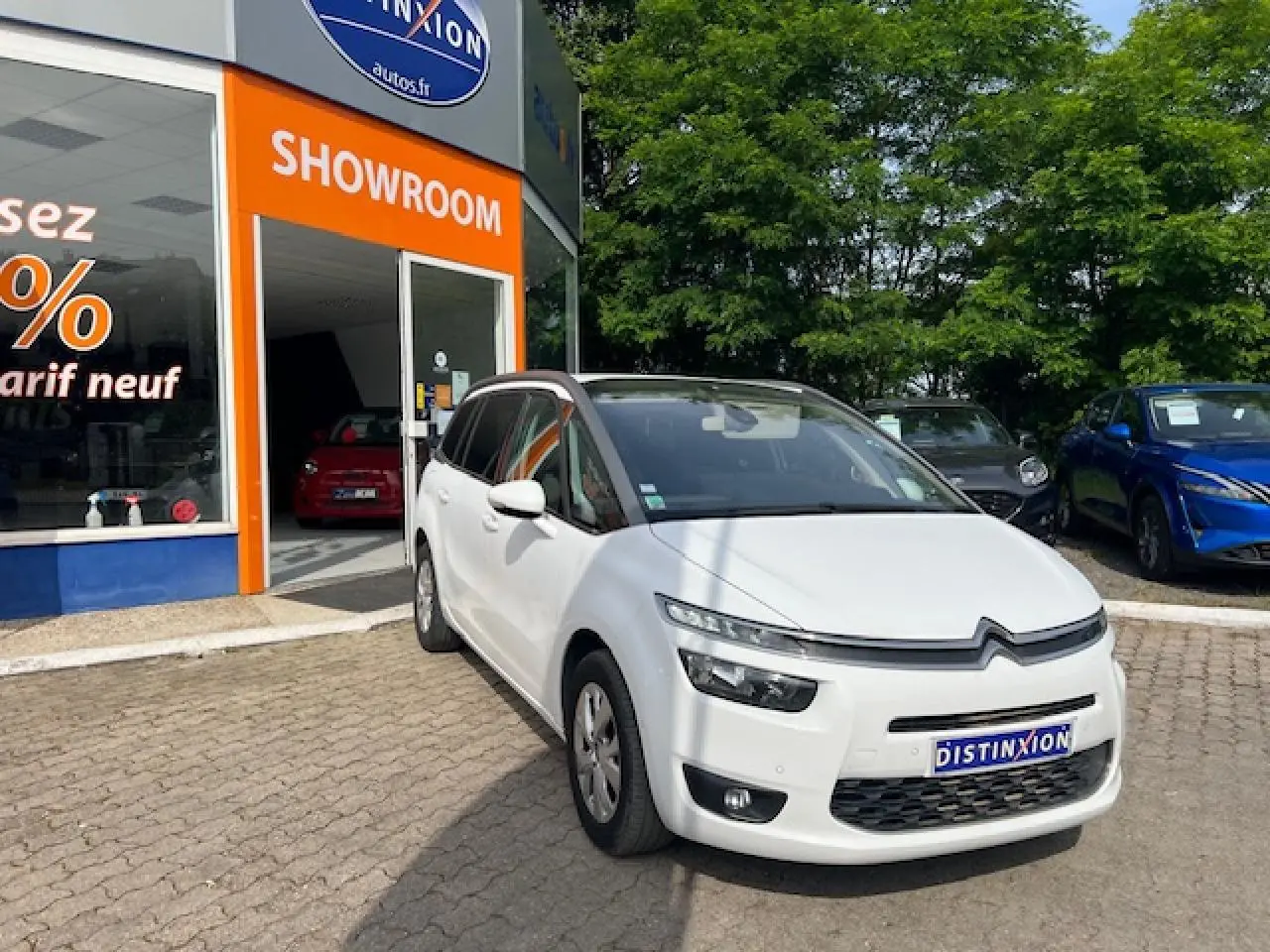 Citroën Grand C4 Picasso blanc en 3/4 avant droit devant un showroom avec végétation en arrière-plan.