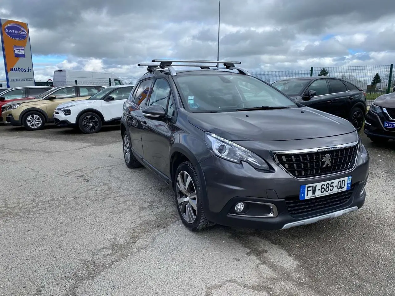 Peugeot 2008 gris platinium vue 3/4 avant droit avec barres de toit aluminium sur parking.
