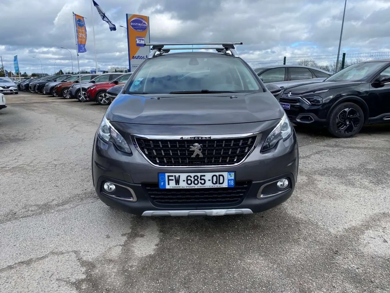 Peugeot 2008 gris platinium vue de face avec barres de toit aluminium sur un parking extérieur.