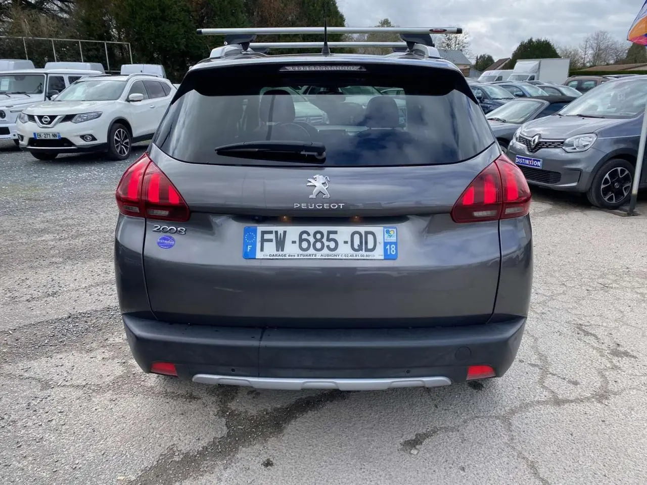 Vue arrière d'un Peugeot 2008 gris platinium avec barres de toit aluminium et caméra de recul visible sur un parking.