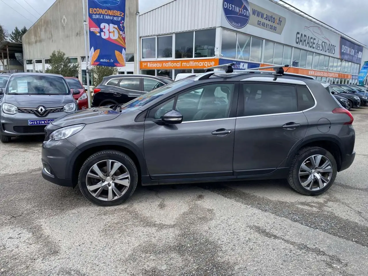 Peugeot 2008 gris platinium vue de profil côté gauche avec barres de toit aluminium sur parking extérieur.