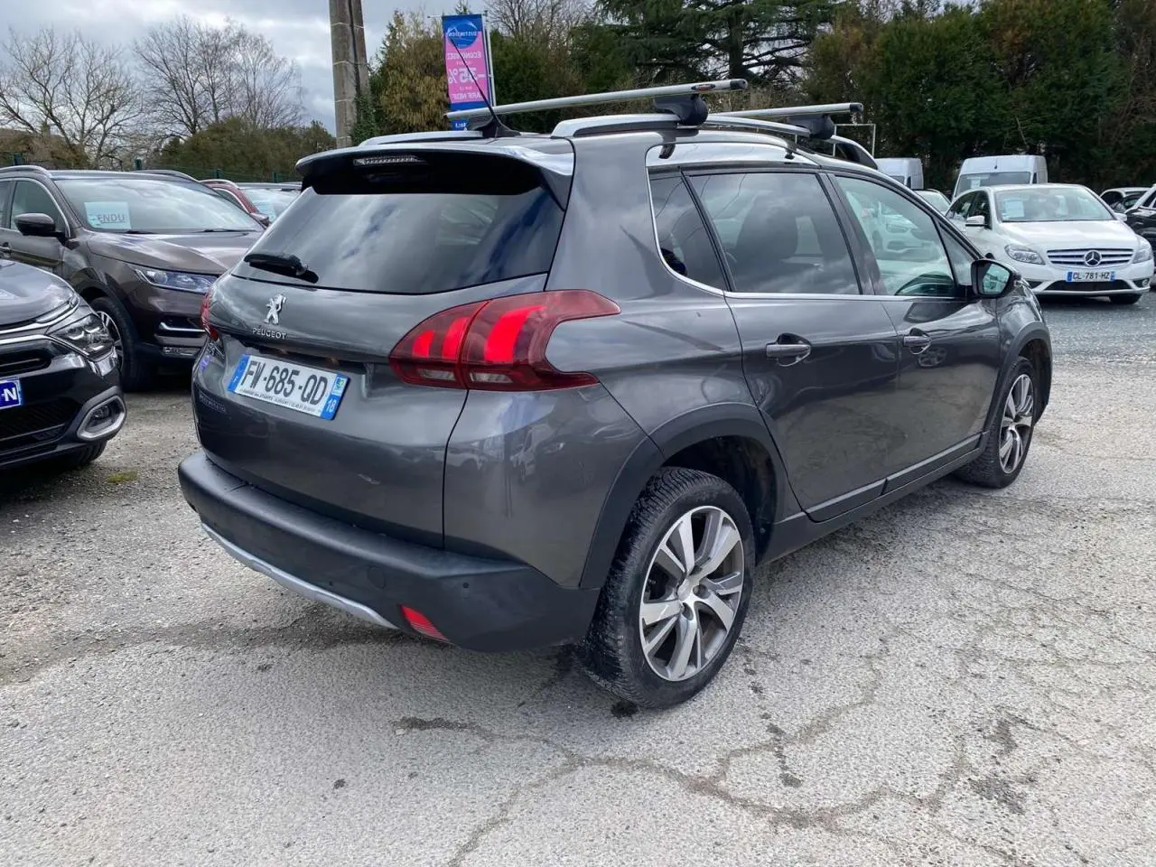 Peugeot 2008 gris platinium vue 3/4 arrière droit avec barres de toit aluminium sur parking extérieur.