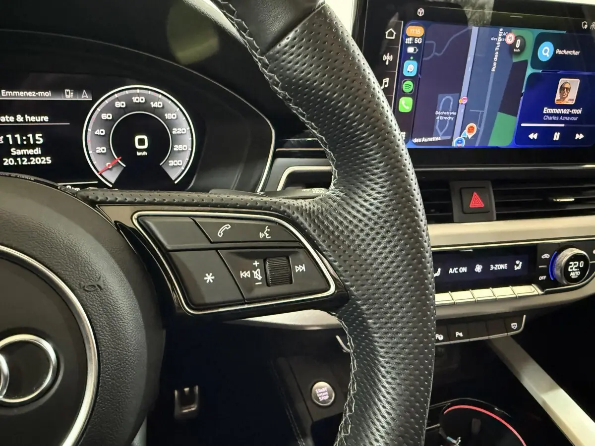 Vue rapprochée de l’intérieur de l’Audi A4 gris foncé, montrant le volant cuir perforé et l’écran tactile central avec navigation.