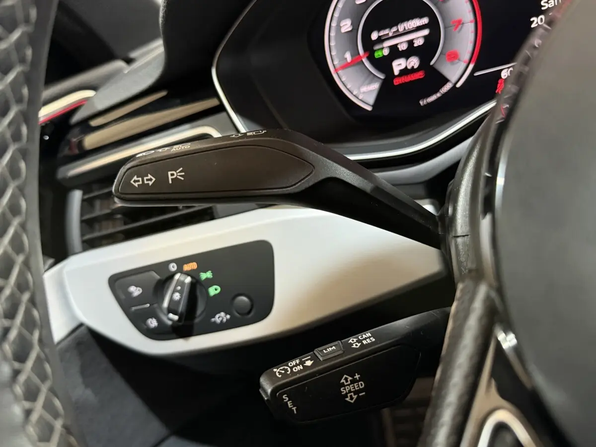 Gros plan sur la commande de clignotants et régulateur de vitesse, tableau de bord numérique partiellement visible, intérieur noir.