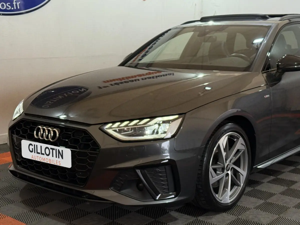 Audi A4 35 TFSI gris foncé vue 3/4 avant droit, avec calandre hexagonale noire et phares LED allumés.