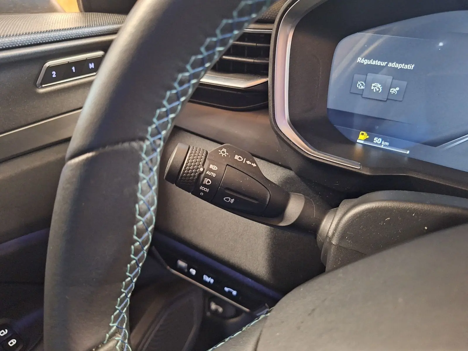 Gros plan sur la commande des phares à gauche du volant avec coutures bleues sur un tableau de bord noir du Lynk & Co 01 hybride 2023.