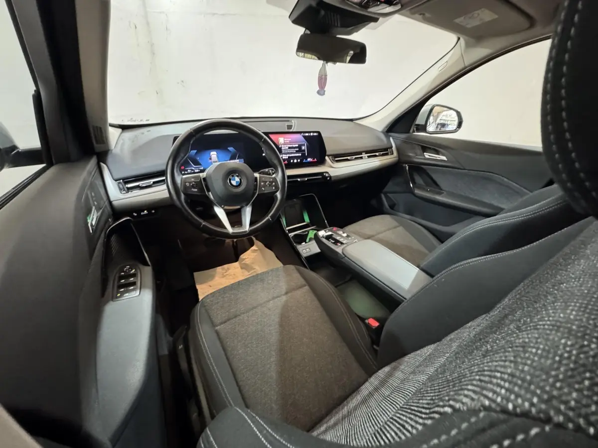 Intérieur BMW X1 2024 vu côté conducteur, sièges gris, volant cuir multifonctions et écran tactile central.